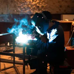 Mistrz pokazuje, jak spawać migomatem, tworząc jasny łuk i niebieski dym podczas pracy nad metalową konstrukcją.