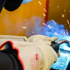 Ręce w rękawicach spawacza pracują nad metalem, iskry i niebieski dym świadczą o trwającym procesie. To praktyczny element kursu spawacza.