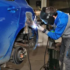 Mechanik w masce spawa blachę samochodową, przywracając jej dawny kształt. Iskry sypią się podczas prac blacharskich.