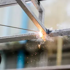 Ręka w rękawicy trzyma elektrodę, wykonując spawanie łukowe metalowych profili. Iskry i dym unoszą się w powietrzu.