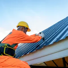 Pracownik w pomarańczowym kombinezonie i żółtym kasku pokazuje, jak wkręcać wkręty do blachy trapezowej, montując dach.