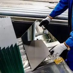 Pracownik w niebieskim kombinezonie i rękawiczkach obraca lustro. Obok stos tafli szkła, które można szlifować.