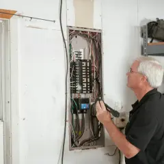 Starszy mężczyzna z latarką bada rozdzielnicę elektryczną. Widoczny jest opis tabliczki znamionowej.