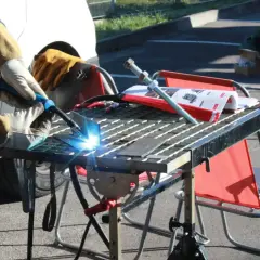 Osoba w rękawicach spawa metal migomatem, tworząc jasny łuk. Na stole leżą instrukcje i śruby. Widać potencjalne błędy przy spawaniu migomatem.