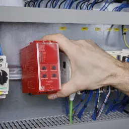 Dłoń instalatora montuje czerwony przekaźnik bezpieczeństwa w szafie sterowniczej, wśród przewodów i innych modułów.
