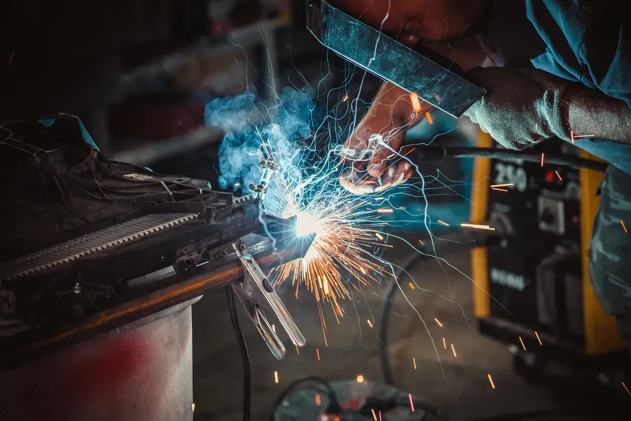Spawacz w masce ochronnej pracuje przy metalowym elemencie, iskry sypią się z miejsca spawania. W tle widać żółtą spawarkę, która może być spawarką inwertorową czy transformatorową.